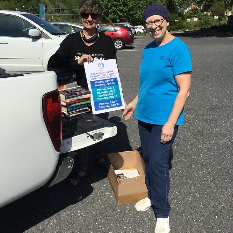 Two volunteers at the tailgate of a pickup truck with a stack of donated books, holding a sign listing donation days